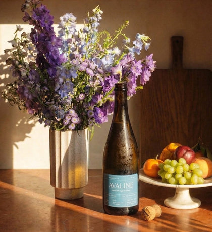 Bottle of Avaline wine with a vase of flowers and fruits on a wooden table.