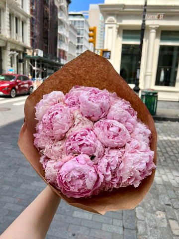 Bouquet of pink flowers wrapped in brown paper held in an urban setting.