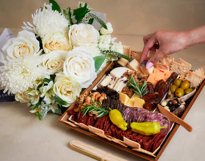 Charcuterie board with meats, cheeses, and olives next to a bouquet of white flowers on a table.