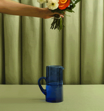 Orange, peach, and cream flower bouquet arranged in a blue glass pitcher on a table against a green curtain background.