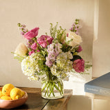 Bouquet of flowers in a vase on a wooden surface with a book and bowl of lemons in the background.
