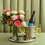 Bouquet of pink roses, bottle of champagne, and two glasses on a tray with a green curtain background.