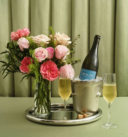 Bouquet of pink roses, bottle of champagne, and two glasses on a tray with a green curtain background.