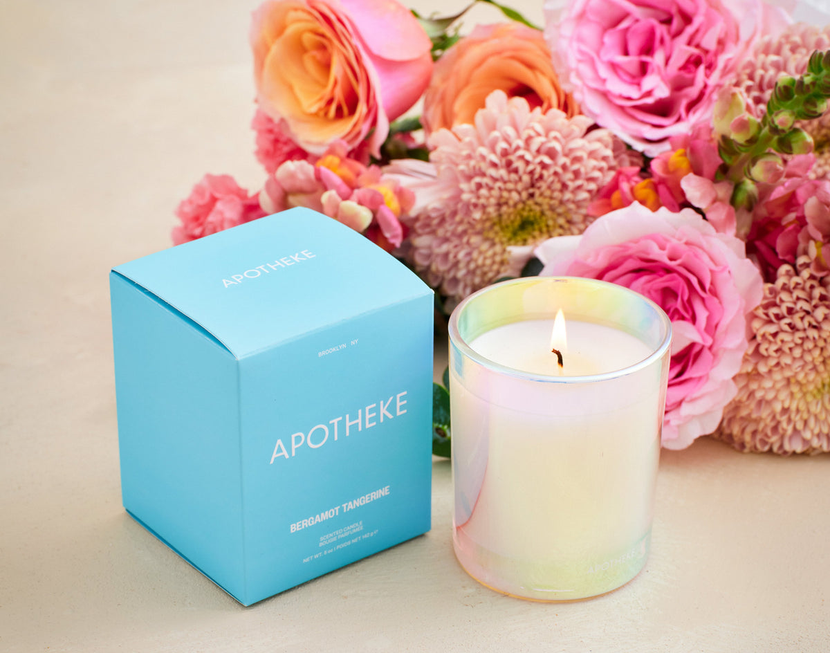 A lit candle with a colorful glass finish next to its blue box, set against a floral background.