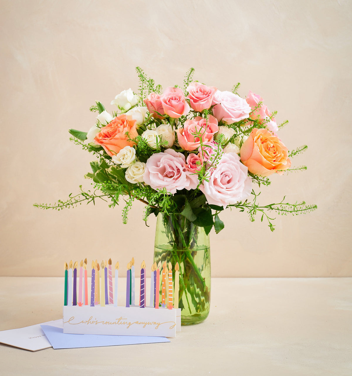 Bouquet of pink and orange roses in a clear vase with birthday candles and card on a beige background