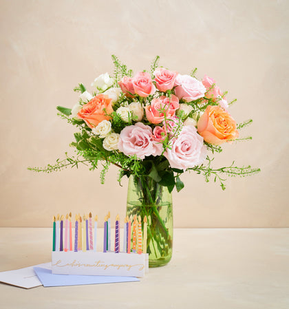 Bouquet of pink and orange roses in a clear vase with birthday candles and card on a beige background