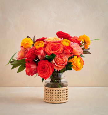 Bouquet of red, orange, and yellow flowers in a clear vase with a wicker base on a beige background