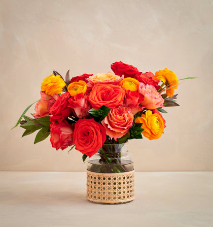 Bouquet of red, orange, and yellow flowers in a clear vase with a wicker base on a beige background